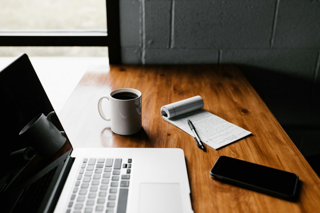 A laptop, a notebook with pen, a mug and a small jar of flowers on a table
